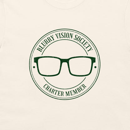 A close-up of a cream-colored t-shirt featuring a dark green circular graphic with the text 'BLURRY VISION SOCIETY CHARTER MEMBER' surrounding an illustration of eyeglasses.