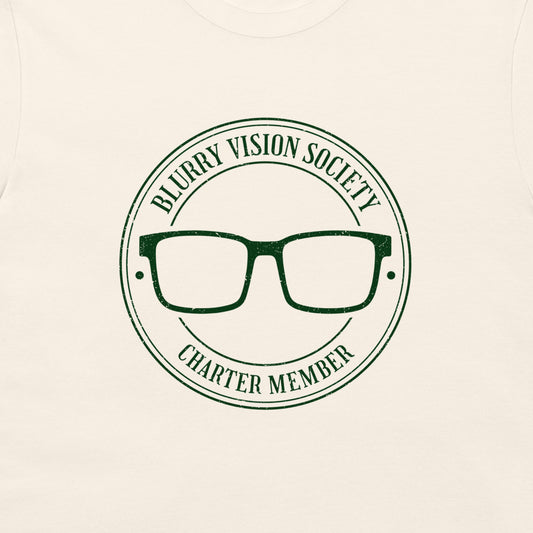 A close-up of a cream-colored t-shirt featuring a dark green circular graphic with the text 'BLURRY VISION SOCIETY CHARTER MEMBER' surrounding an illustration of eyeglasses.