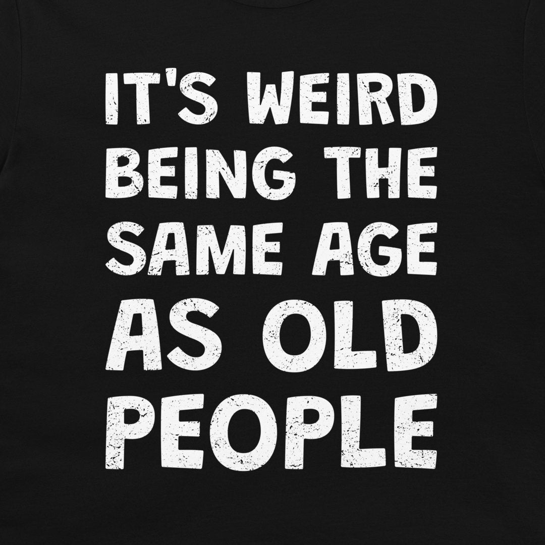 A close-up of a black garment, likely a t-shirt, featuring a distressed white typographic print that reads 'IT'S WEIRD BEING THE SAME AGE AS OLD PEOPLE'.