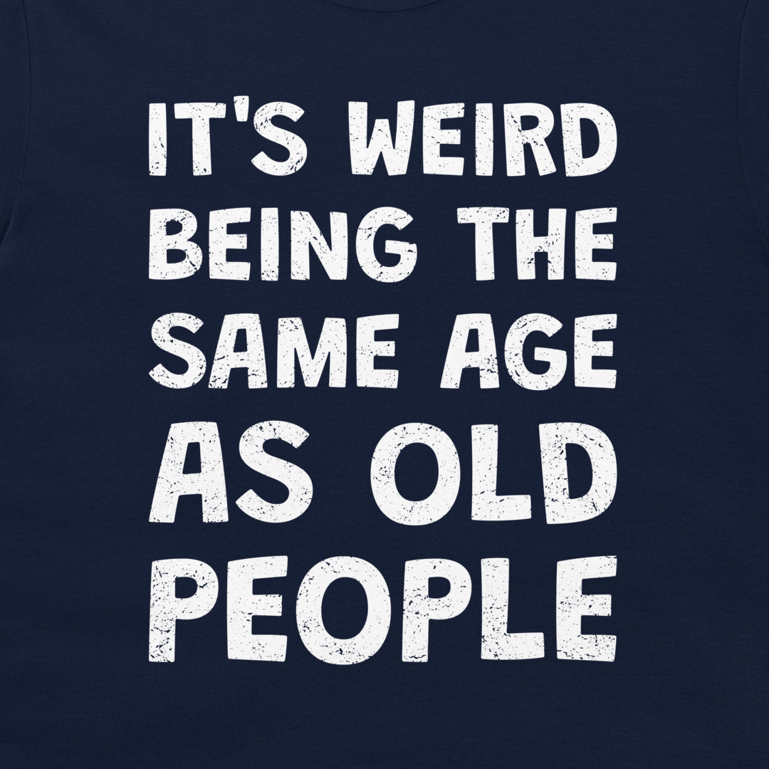 A close-up of a navy blue t-shirt featuring a bold, distressed white text graphic that reads 'IT'S WEIRD BEING THE SAME AGE AS OLD PEOPLE'.