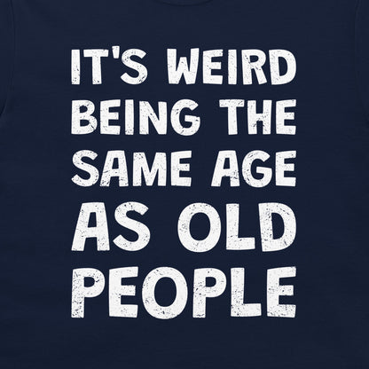 A close-up of a navy blue t-shirt featuring a bold, distressed white text graphic that reads 'IT'S WEIRD BEING THE SAME AGE AS OLD PEOPLE'.