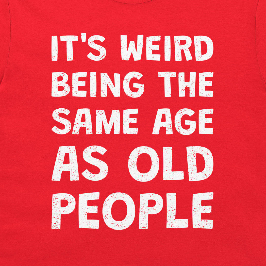 A close-up of a bright red t-shirt, featuring a bold white distressed text graphic that reads 'IT'S WEIRD BEING THE SAME AGE AS OLD PEOPLE'.