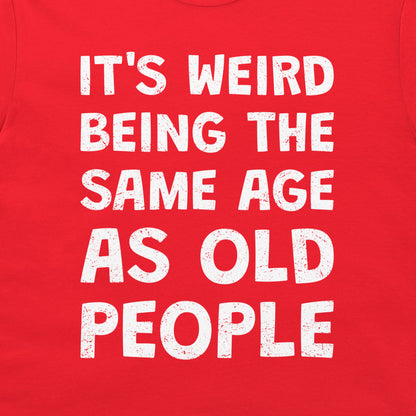 A close-up of a bright red t-shirt, featuring a bold white distressed text graphic that reads 'IT'S WEIRD BEING THE SAME AGE AS OLD PEOPLE'.