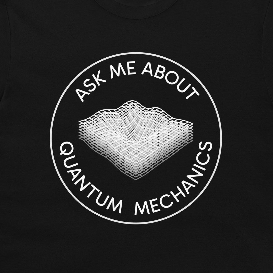 A close-up of a black t-shirt, featuring a white circular graphic with the text 'ASK ME ABOUT QUANTUM MECHANICS' surrounding a white wireframe illustration of a wave-particle duality concept.