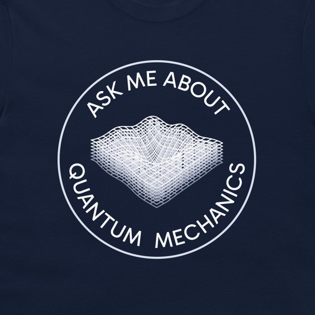 A close-up of a navy blue t-shirt, featuring a white circular graphic with the text 'ASK ME ABOUT QUANTUM MECHANICS' surrounding a wireframe 3D landscape illustration.