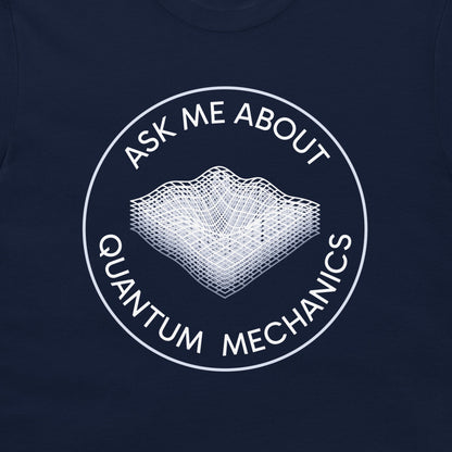 A close-up of a navy blue t-shirt, featuring a white circular graphic with the text 'ASK ME ABOUT QUANTUM MECHANICS' surrounding a wireframe 3D landscape illustration.