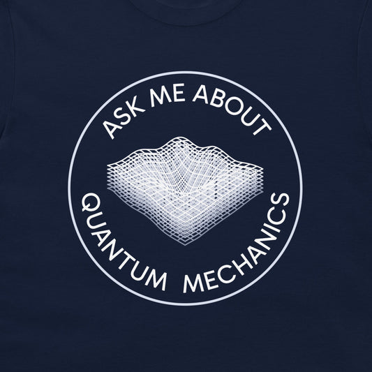A close-up of a navy blue t-shirt, featuring a white circular graphic with the text 'ASK ME ABOUT QUANTUM MECHANICS' surrounding a wireframe 3D landscape illustration.