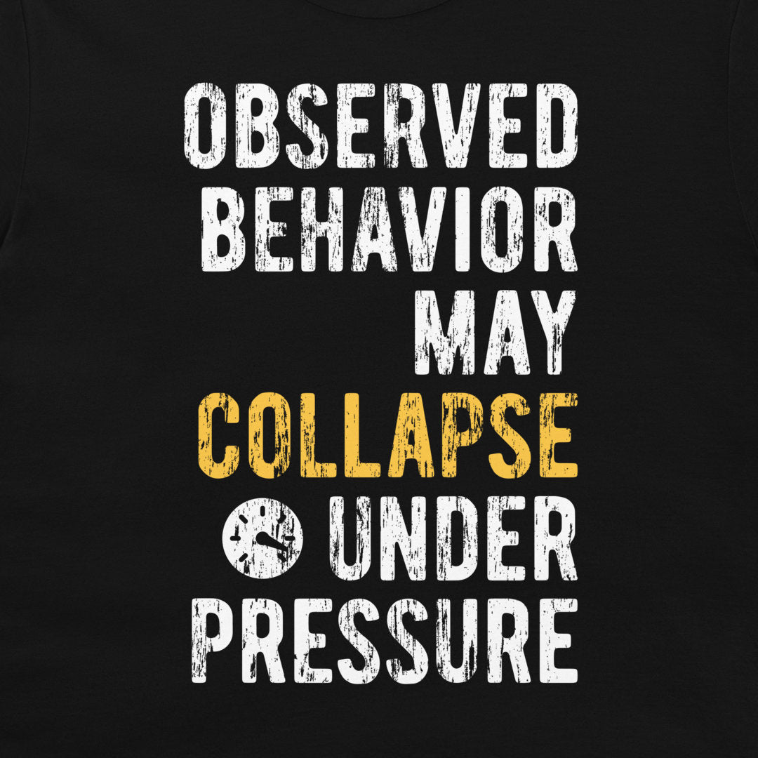 A close-up of a black graphic t-shirt featuring a distressed typographic design that reads 'OBSERVED BEHAVIOR MAY COLLAPSE UNDER PRESSURE' in white and yellow lettering.
