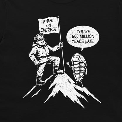 A black graphic print on a t-shirt featuring a white cartoon illustration of a climber reaching the top of Everest with a flag that reads 'FIRST ON EVEREST', next to a trilobite with a speech bubble saying 'YOU'RE 500 MILLION YEARS LATE'.