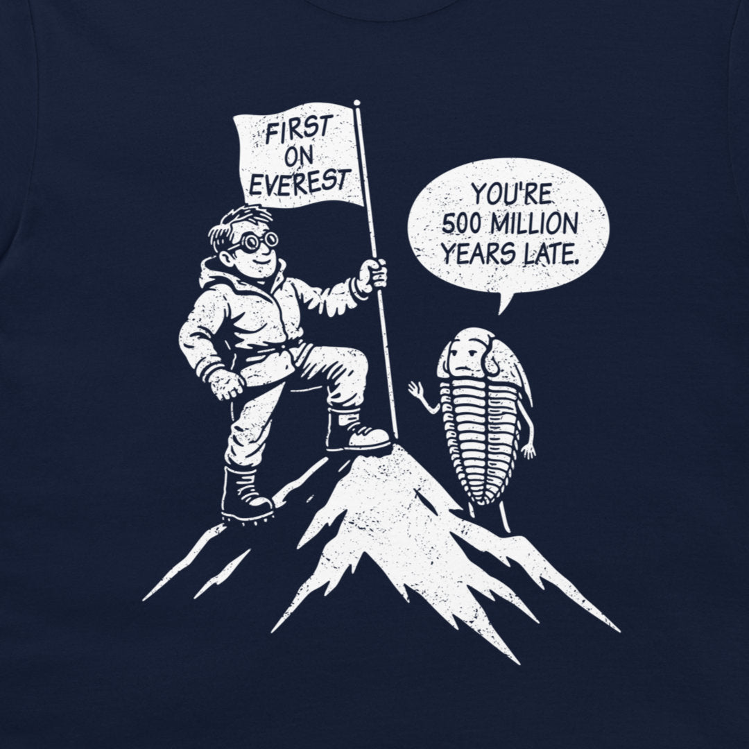 A navy blue graphic print on a t-shirt featuring a white cartoon illustration of a climber reaching the top of Everest with a flag that reads 'FIRST ON EVEREST', next to a trilobite with a speech bubble saying 'YOU'RE 500 MILLION YEARS LATE'.