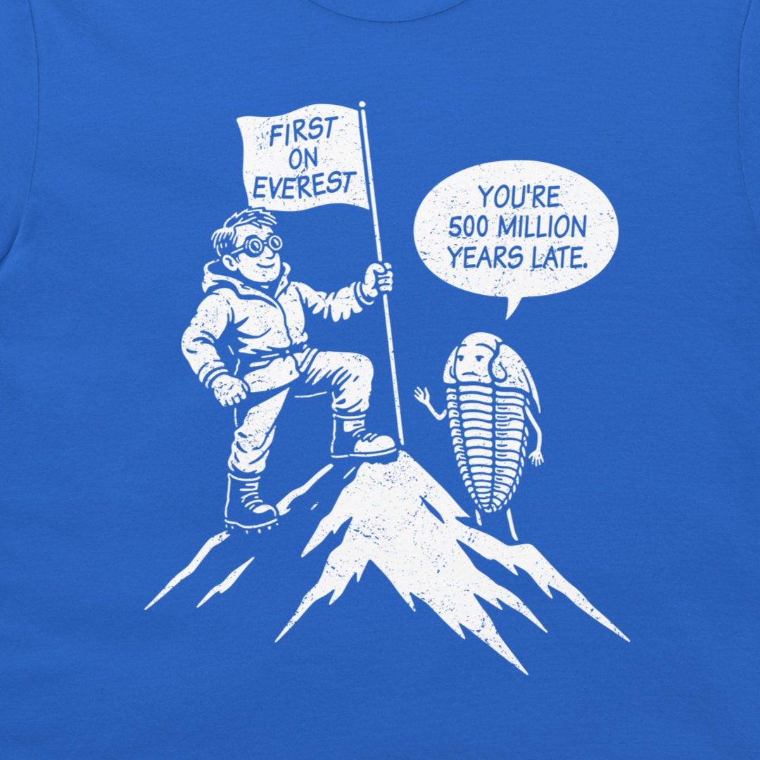 A close-up of a royal blue graphic print on a t-shirt featuring a white cartoon illustration of a climber reaching the top of Everest with a flag that reads 'FIRST ON EVEREST', next to a trilobite with a speech bubble saying 'YOU'RE 500 MILLION YEARS LATE'.