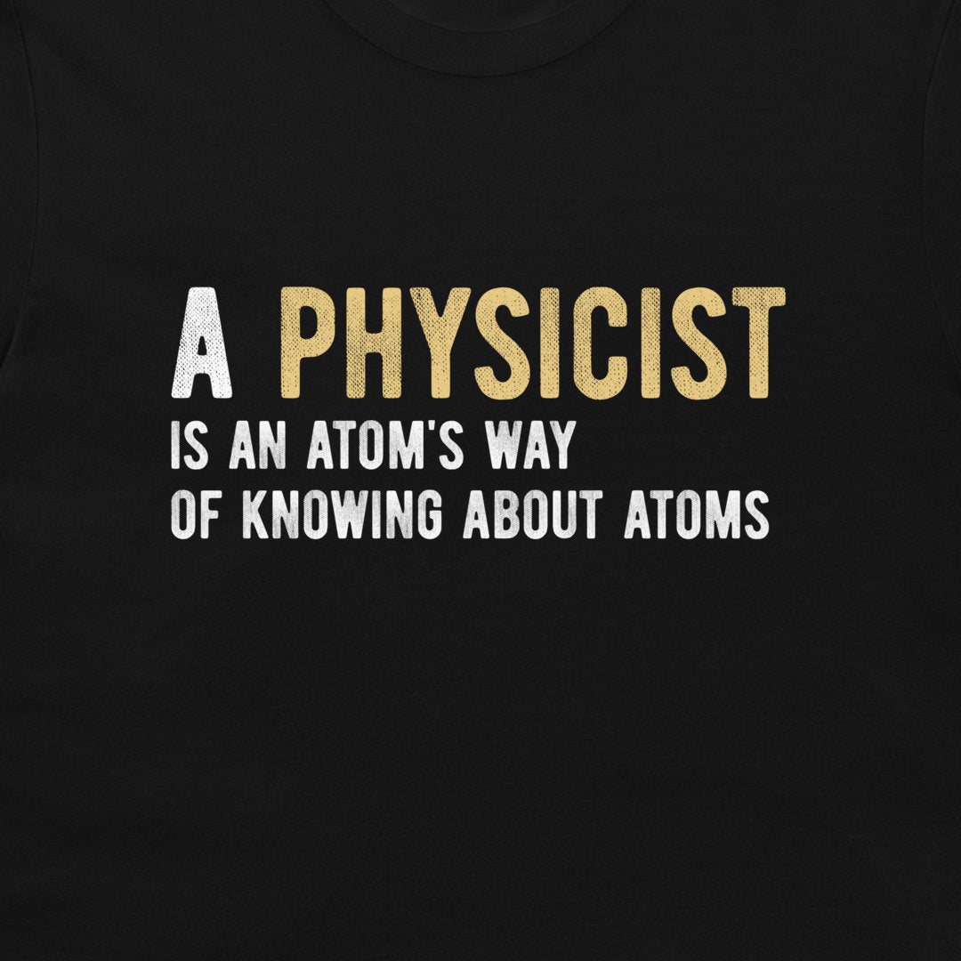 A close-up of a black t-shirt featuring a printed graphic with the text 'A PHYSICIST IS AN ATOM'S WAY OF KNOWING ABOUT ATOMS' in white and gold lettering.