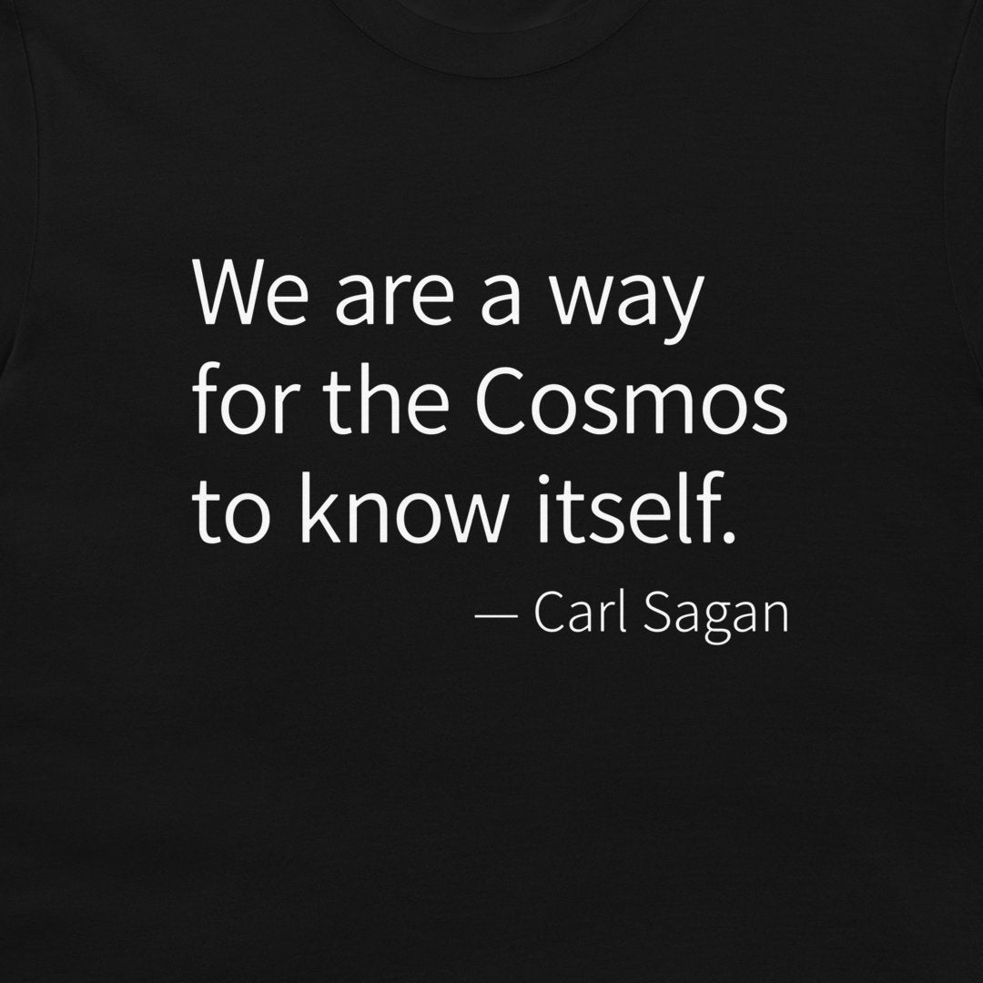 A close-up of a black t-shirt, featuring a white printed quote by Carl Sagan: 'We are a way for the Cosmos to know itself.'.
