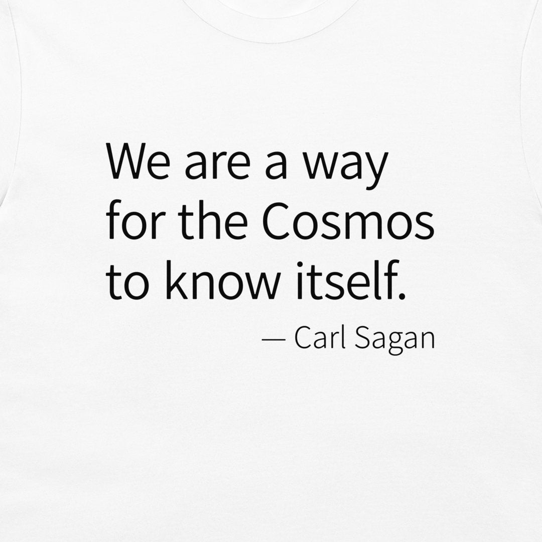 A close-up of a white T-shirt, printed with the quote 'We are a way for the Cosmos to know itself.' and '— Carl Sagan' in black serif text.