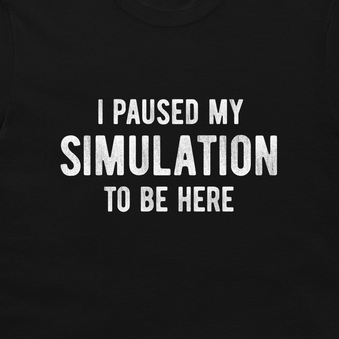 A close-up of a black t-shirt featuring the phrase 'I PAUSED MY SIMULATION TO BE HERE' printed in distressed white capital letters.