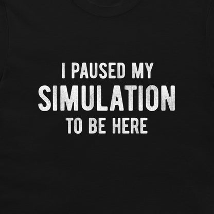 A close-up of a black t-shirt featuring the phrase 'I PAUSED MY SIMULATION TO BE HERE' printed in distressed white capital letters.