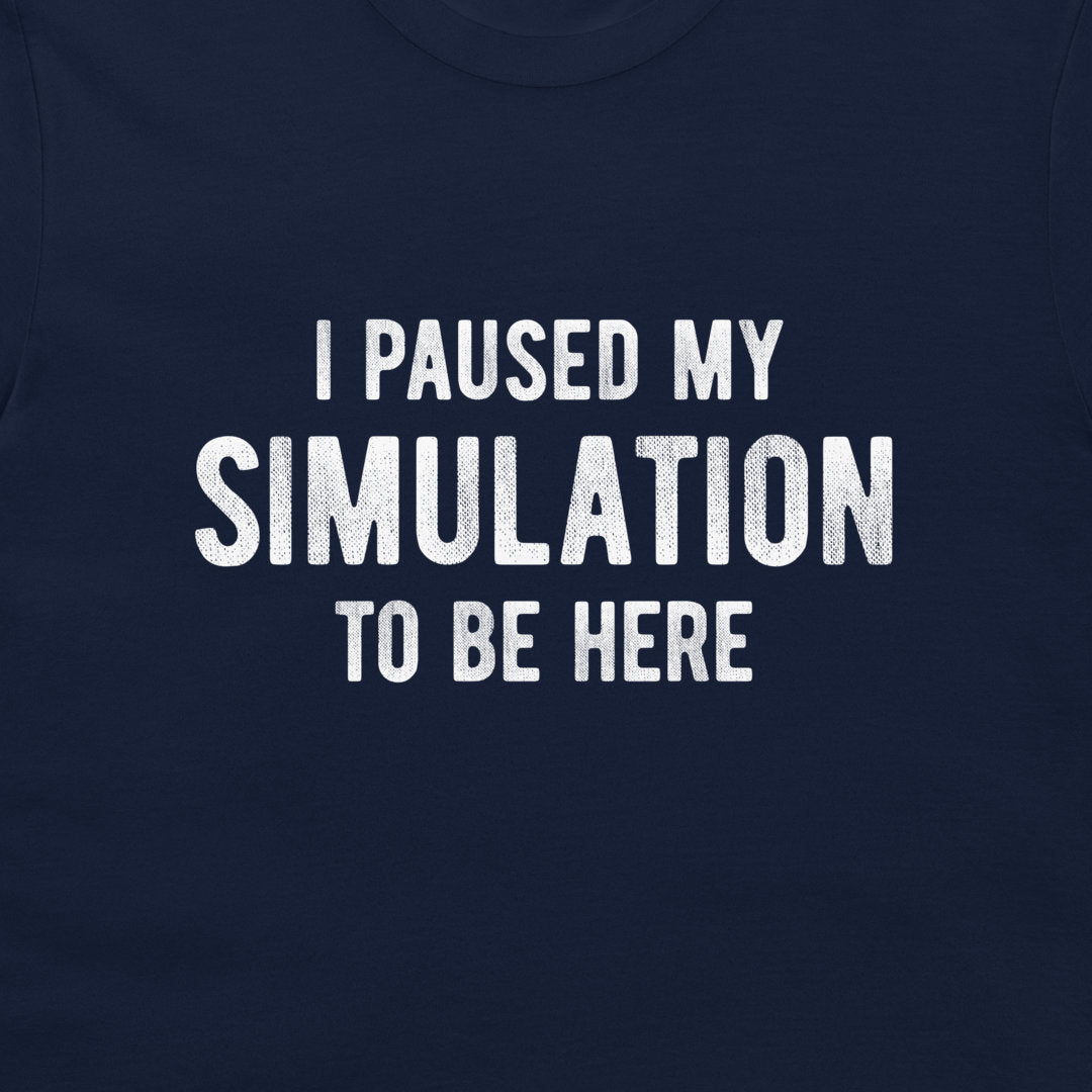A close-up of a navy blue t-shirt featuring the phrase 'I PAUSED MY SIMULATION TO BE HERE' printed in distressed white capital letters across the chest.