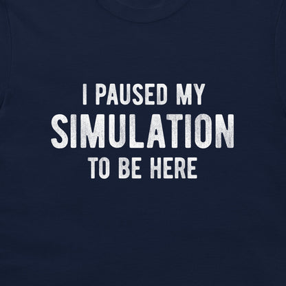 A close-up of a navy blue t-shirt featuring the phrase 'I PAUSED MY SIMULATION TO BE HERE' printed in distressed white capital letters across the chest.