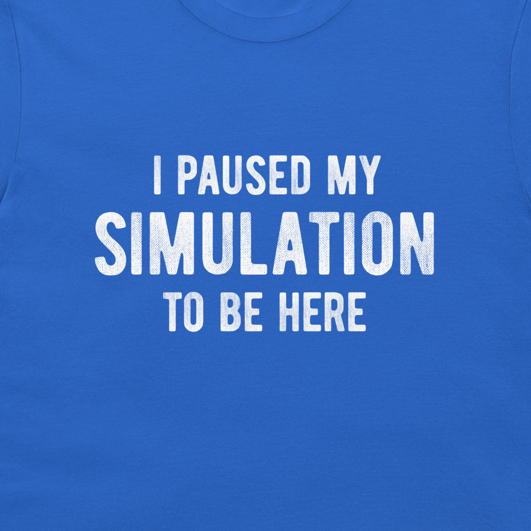 A close-up of a royal blue t-shirt featuring the phrase 'I PAUSED MY SIMULATION TO BE HERE' printed in distressed white capital letters across the chest.