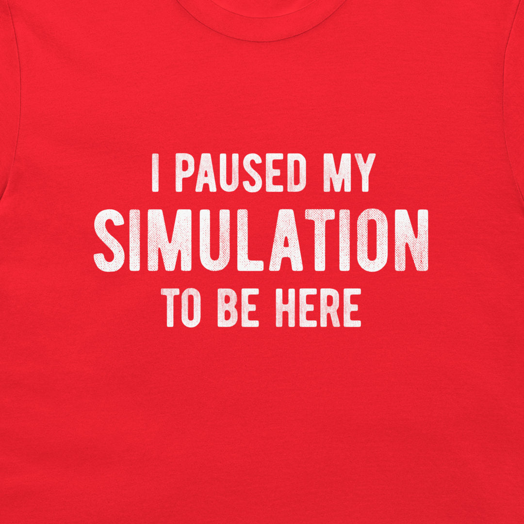 A close-up of a bright red t-shirt featuring the phrase 'I PAUSED MY SIMULATION TO BE HERE' printed in distressed white capital letters across the chest.