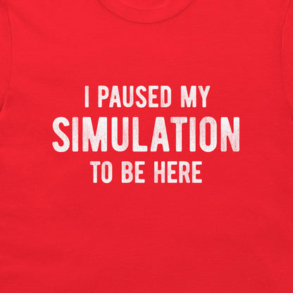 A close-up of a bright red t-shirt featuring the phrase 'I PAUSED MY SIMULATION TO BE HERE' printed in distressed white capital letters across the chest.