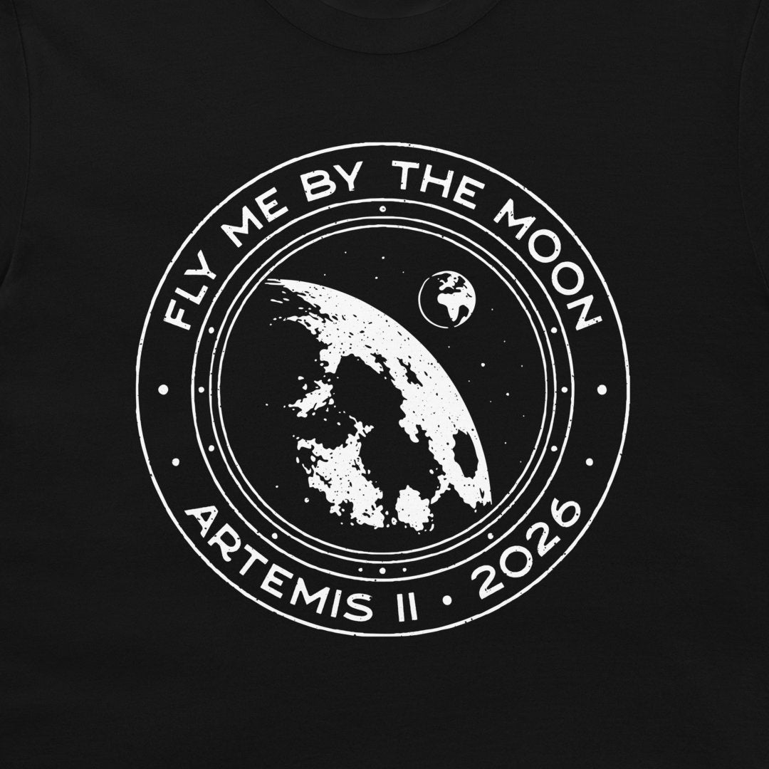 A close-up of a black t-shirt, featuring a distressed white circular graphic print with the text 'FLY ME BY THE MOON', 'ARTEMIS II', and '2026' surrounding an illustration of the Earth and Moon.