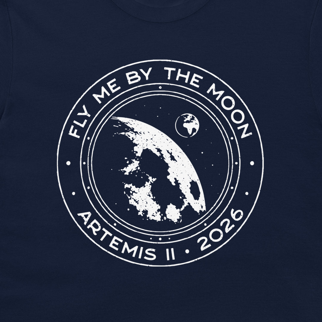A close-up of a navy blue t-shirt, featuring a distressed white circular graphic print with the text 'FLY ME BY THE MOON', 'ARTEMIS II', and '2026' surrounding an illustration of the Earth and Moon.