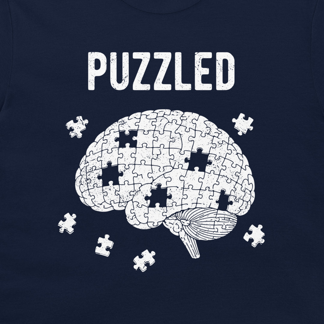 A navy blue t-shirt featuring a white graphic of a brain made of puzzle pieces with several missing pieces scattered around it, and the word 'PUZZLED' printed above in a distressed white font.