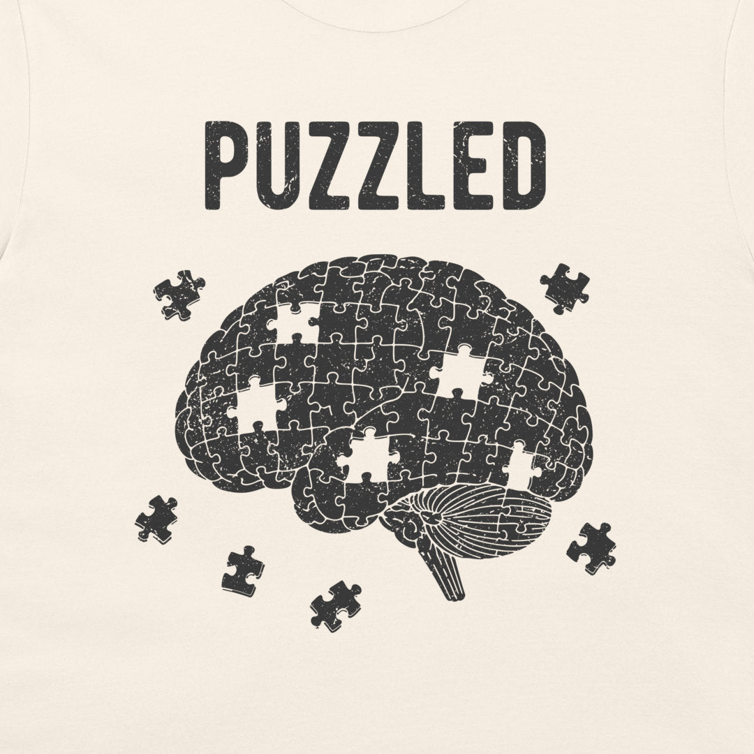 A close-up of a light cream-colored garment, likely a t-shirt, featuring a black graphic print of a brain made of puzzle pieces with the word 'PUZZLED' above it and a few loose puzzle pieces around it.