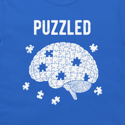 A close-up of a blue T-shirt or sweatshirt featuring a white graphic of a brain made of puzzle pieces, with the word 'PUZZLED' printed above and a few loose puzzle pieces around it.