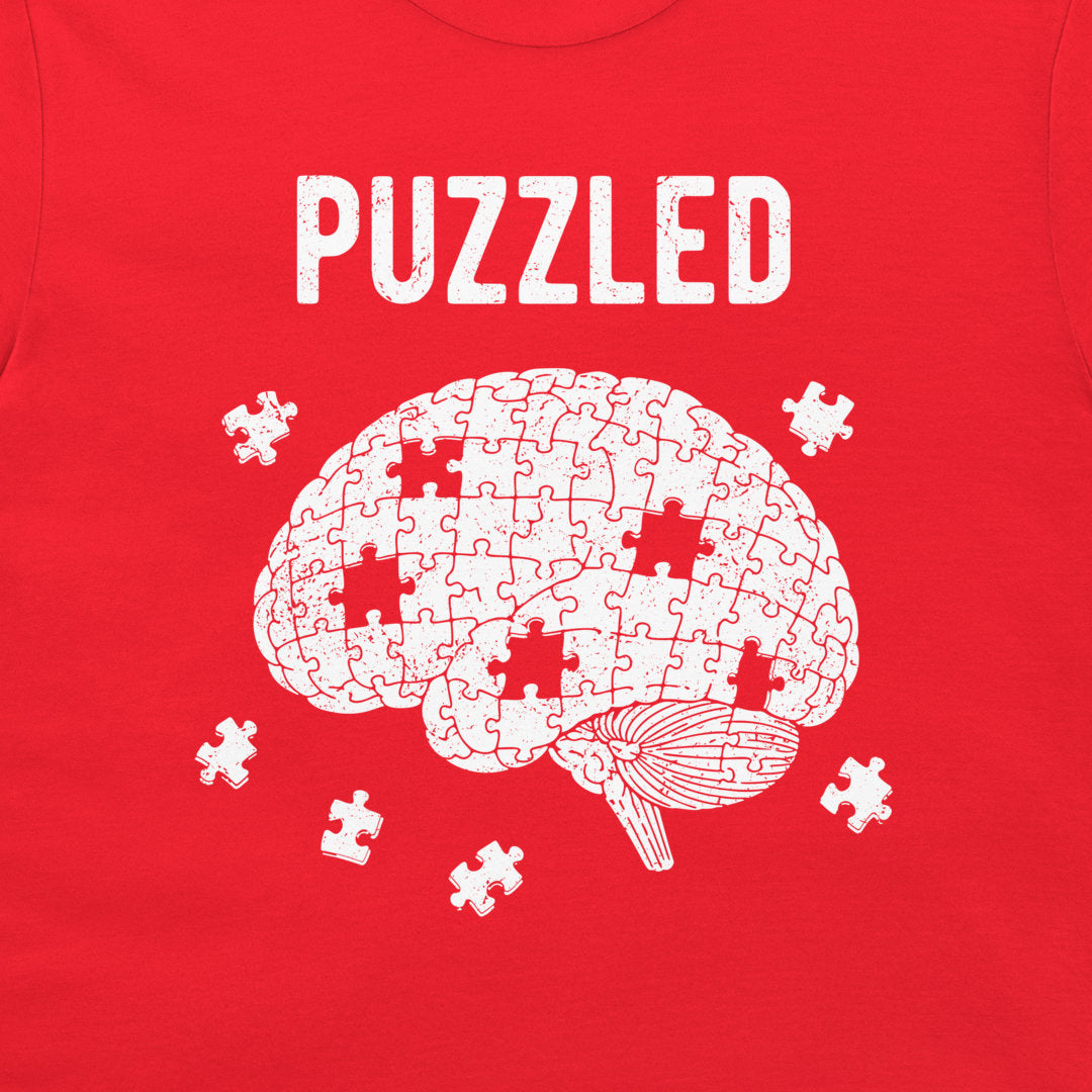 A red t-shirt featuring a white graphic of a brain made of puzzle pieces with several missing pieces scattered around it, and the word 'PUZZLED' printed above in a distressed white font.