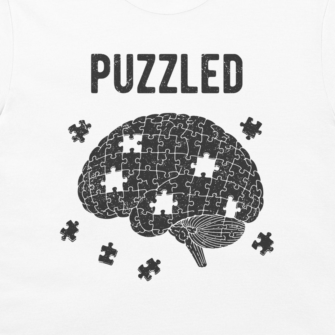 A white t-shirt featuring a black graphic print of a brain made of jigsaw puzzle pieces, with the word 'PUZZLED' printed above and several loose puzzle pieces scattered around it.