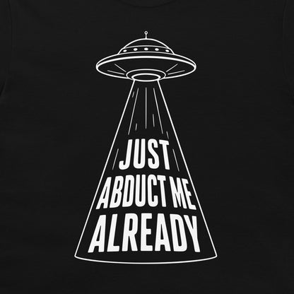 A black t-shirt featuring a white graphic of a classic flying saucer projecting a spotlight with the text 'JUST ABDUCT ME ALREADY' inside the beam.