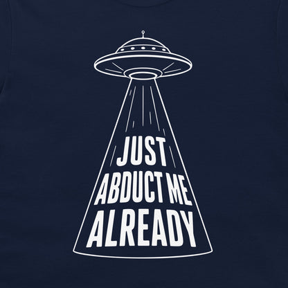 A close-up of a navy blue t-shirt, featuring a white graphic of a UFO with a beam of light projecting the text 'JUST ABDUCT ME ALREADY'.