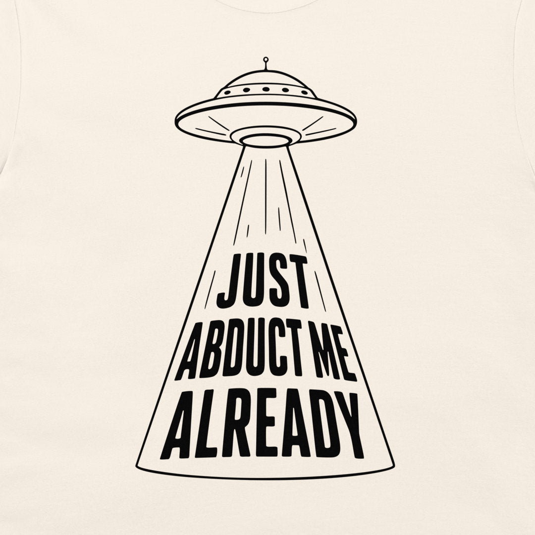 A close-up of a light beige or off-white fabric surface featuring a black line-art graphic of a UFO with a spotlight beam and the text 'JUST ABDUCT ME ALREADY'.