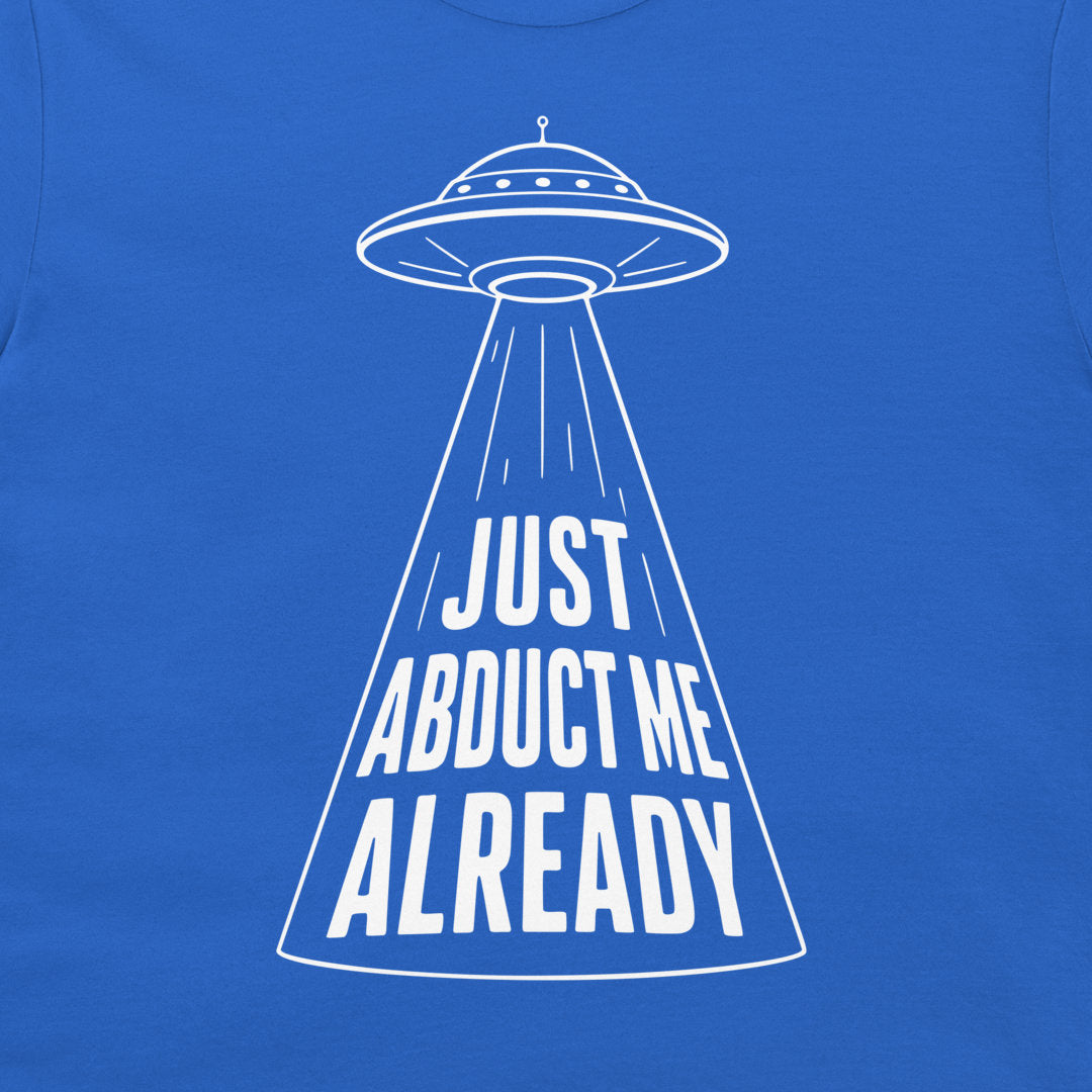 A close-up of a royal blue t-shirt featuring a white graphic of a flying saucer projecting the text 'JUST ABDUCT ME ALREADY' in a bold, stylized font.