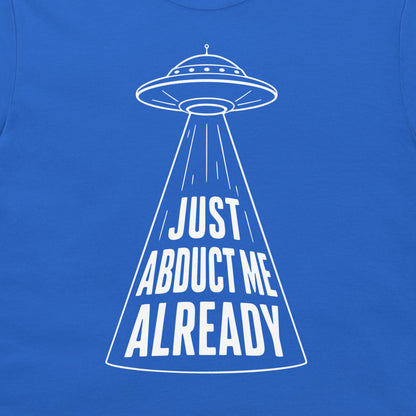 A close-up of a royal blue t-shirt featuring a white graphic of a flying saucer projecting the text 'JUST ABDUCT ME ALREADY' in a bold, stylized font.