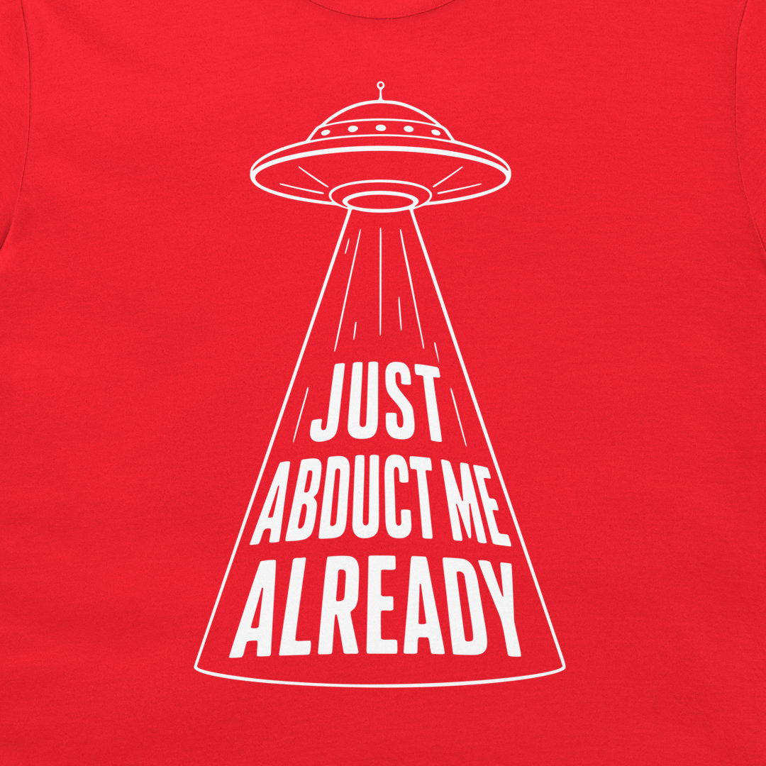 A close-up of a bright red t-shirt, featuring a white line-art graphic of a UFO with a spotlight beam and the text 'JUST ABDUCT ME ALREADY' printed inside the beam.