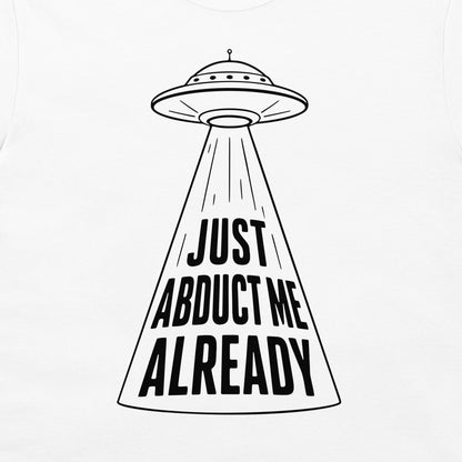 A white t-shirt featuring a black graphic of a classic flying saucer UFO casting a beam of light with the bold text 'JUST ABDUCT ME ALREADY'.