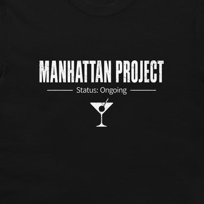 A close-up of a black t-shirt, featuring a white distressed graphic print that reads 'MANHATTAN PROJECT' with 'Status: Ongoing' and a martini glass icon below.