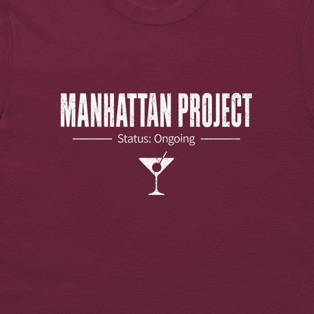 A close-up of a maroon crewneck t-shirt featuring a white distressed graphic that reads 'MANHATTAN PROJECT' with 'Status: Ongoing' and a martini glass icon below.