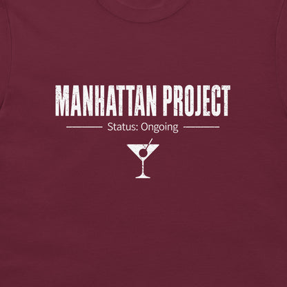 A close-up of a maroon crewneck t-shirt featuring a white distressed graphic that reads 'MANHATTAN PROJECT' with 'Status: Ongoing' and a martini glass icon below.