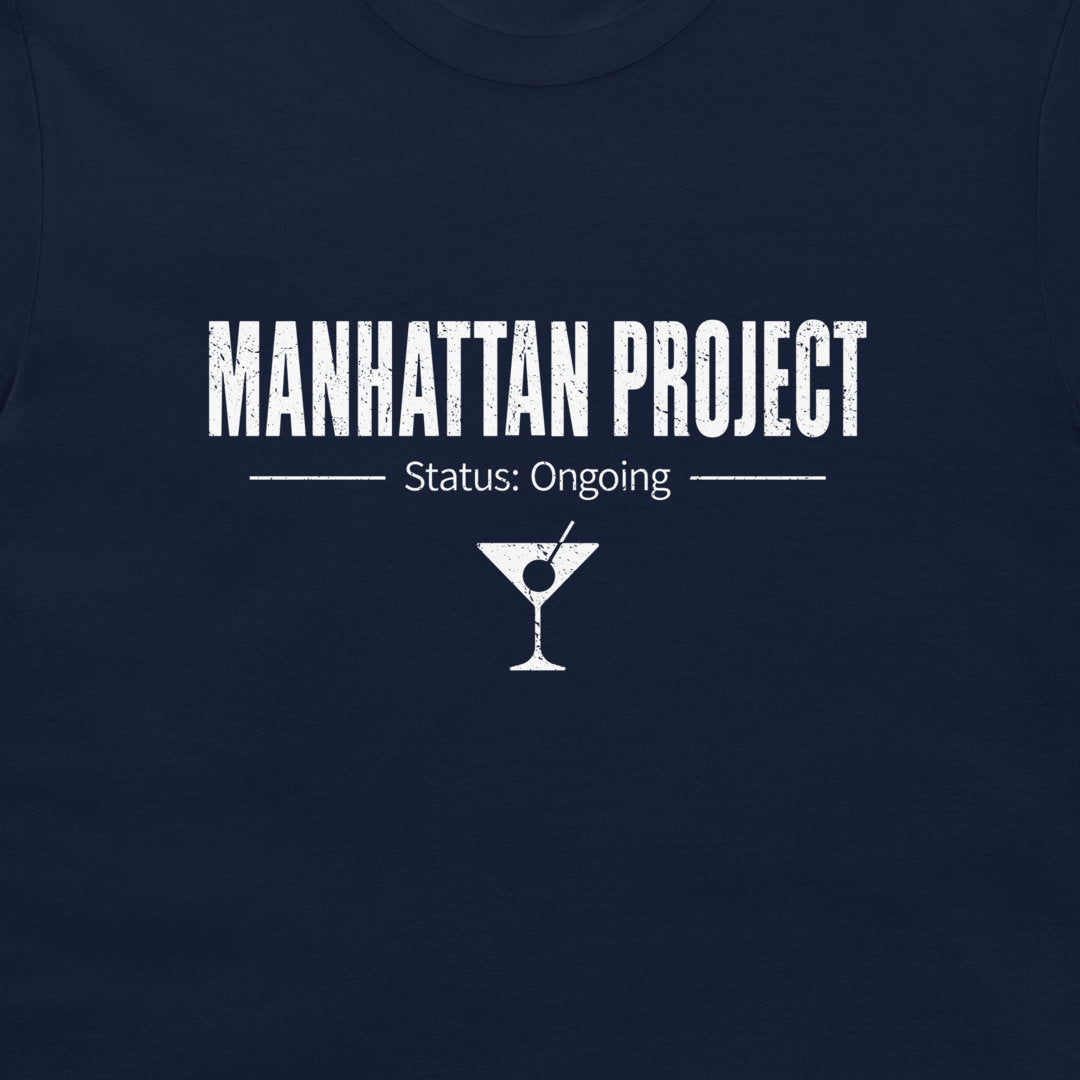 A close-up of a navy blue t-shirt featuring a white graphic print that reads 'MANHATTAN PROJECT' with 'Status: Ongoing' and a martini glass icon below.