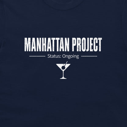 A close-up of a navy blue t-shirt featuring a white graphic print that reads 'MANHATTAN PROJECT' with 'Status: Ongoing' and a martini glass icon below.