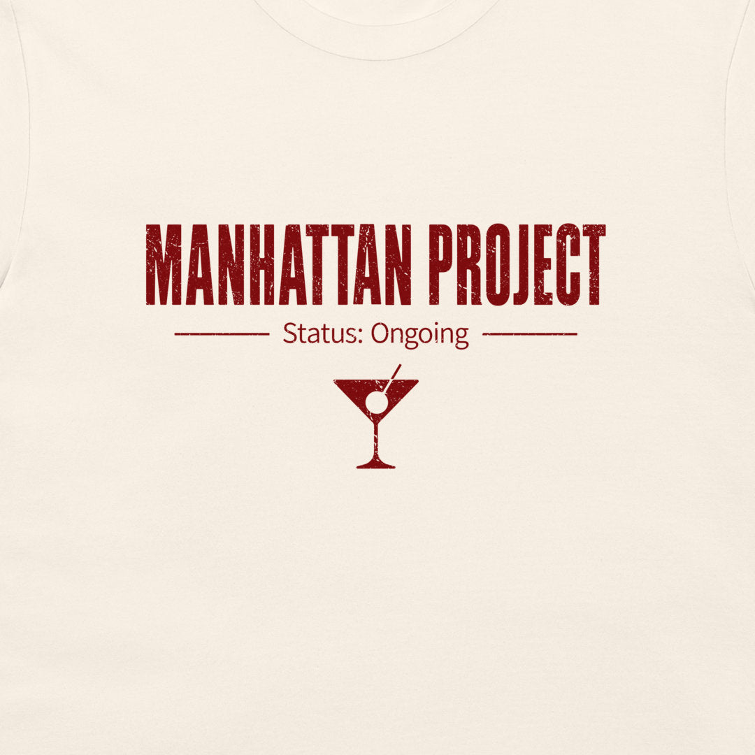 A close-up of a light cream-colored t-shirt, featuring a dark red graphic print that reads 'MANHATTAN PROJECT' and 'Status: Ongoing' with a martini glass icon.