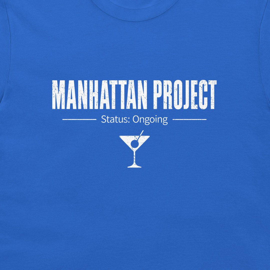 A close-up of a royal blue t-shirt featuring a white distressed graphic print that reads 'MANHATTAN PROJECT' with 'Status: Ongoing' and a martini glass icon below.