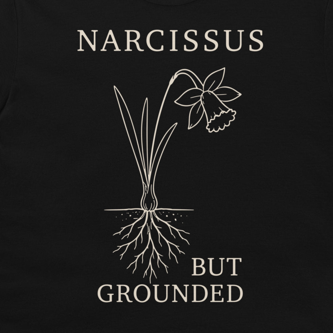 A close-up of a black t-shirt, featuring a white graphic print of a daffodil with its roots exposed and the text 'NARCISSUS BUT GROUNDED' in a serif font.