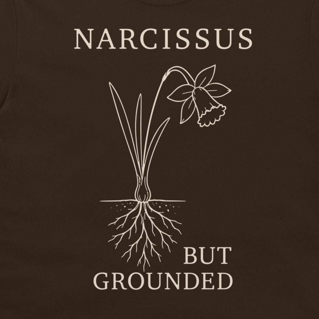 A close-up of a dark brown t-shirt featuring a white graphic of a narcissus flower with its roots exposed, accompanied by the text 'NARCISSUS BUT GROUNDED' in a white serif font.