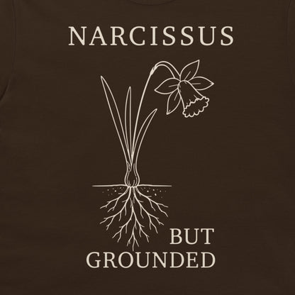 A close-up of a dark brown t-shirt featuring a white graphic of a narcissus flower with its roots exposed, accompanied by the text 'NARCISSUS BUT GROUNDED' in a white serif font.