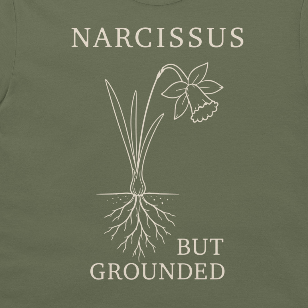 Close-up of an olive green t-shirt featuring a white line art graphic of a daffodil with its roots exposed, accompanied by the text 'NARCISSUS BUT GROUNDED' in a matching off-white serif font.